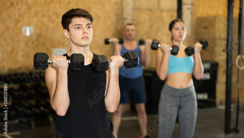 Sportive strong young male in activewear lifting dumbbells during group workout class in gym indoors. Functional training concept © JackF