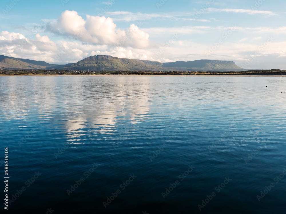Benbulben mountain and blue water surface of Atlantic ocean with reflection of blue cloudy sky. Irish nature. County Sligo, Ireland. Popular tourist attraction. Stunning scenery.