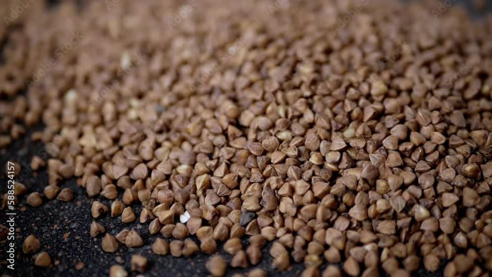 Grains of Buckwheat Fall on a Black Background in Slow Motion. Close up. Raw brown granules and buckwheat particles pour into a pile of buckwheat. Falling buckwheat fills a grainy surface. Texture. Grains of Buckwheat Fall on a Black Background in Slow Motion. Close up. Raw brown granules and buckwheat particles pour into a pile of buckwheat. Falling buckwheat fills a grainy surface. Texture.