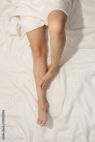 Female legs in bed. Pretty woman lying barefoot in seductive position showing her shapely female legs and feet with french pedicure. Girl is relaxing in white linen bedding. Relax time concept.