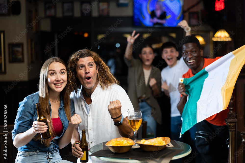 Excited young adult couple of football fans sitting at table in sports bar, watching match together and emotionally cheering for favorite team against background of people with Irish flag