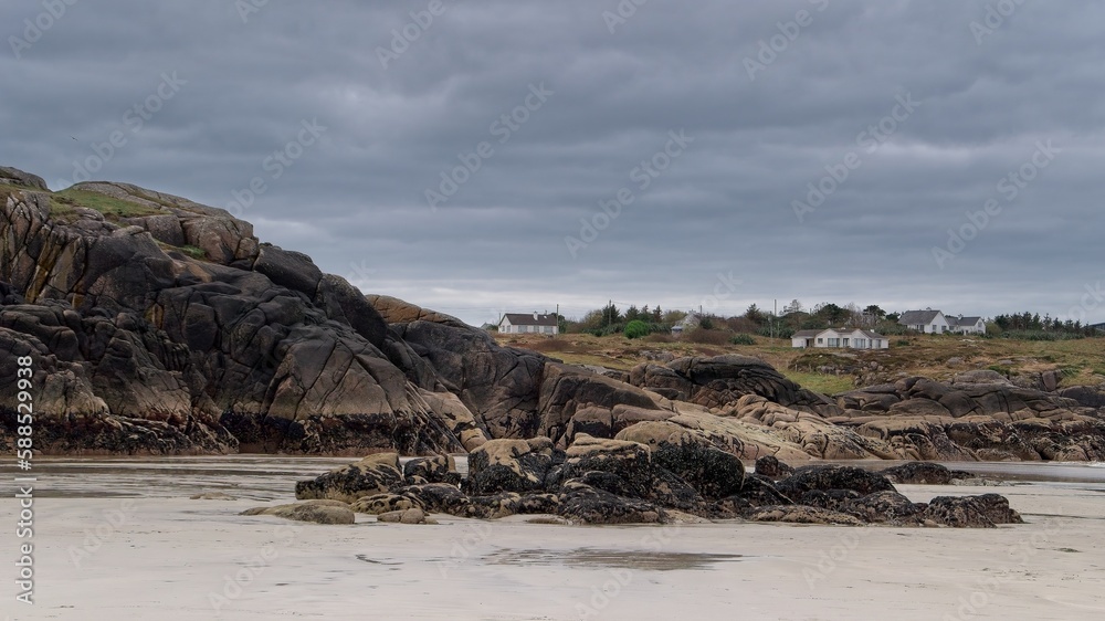 Rocks, cliffs and beach on ocean coast, farm houses on background, dark gray sky, Donegal, Ireland