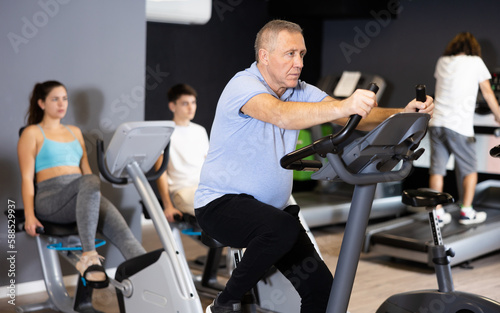 Fototapeta Naklejka Na Ścianę i Meble -  Portrait of active elderly man doing cardio training, cycling on stationary bike in gym ..