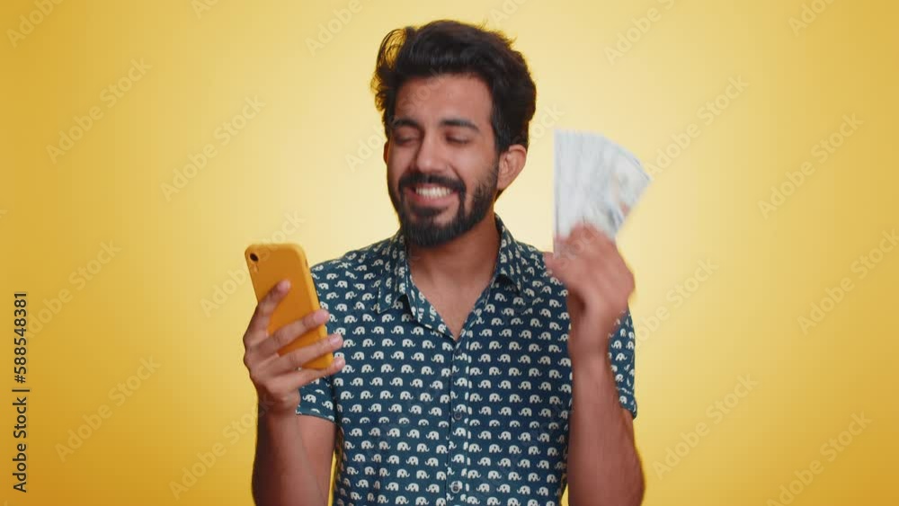Indian man looking smartphone display sincerely rejoicing win, receiving money dollar cash banknotes, success lottery jackpot luck, income salary wage. Hindu guy isolated on studio yellow background Indian man looking smartphone display sincerely rejoicing win, receiving money dollar cash banknotes, success lottery jackpot luck, income salary wage. Hindu guy isolated on studio yellow background