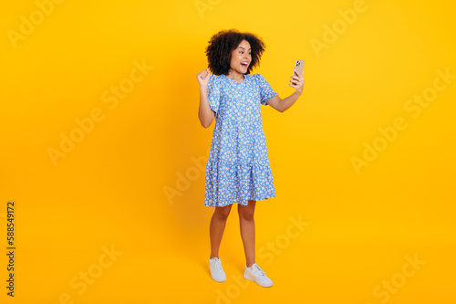 Full-length cheerful african american or brazilian curly woman in summer dress, holds smart phone in hand, making gesture yes, rejoicing in success, victory, news, stand on isolated orange background