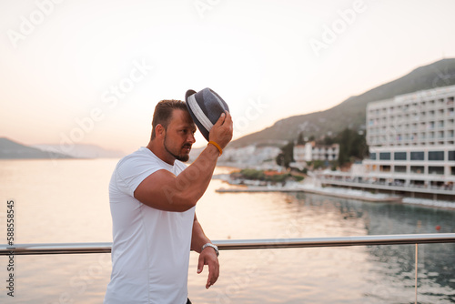 A fit and attractive man on vacation at a hotel near a sandy beach. He is wearing a stylish hat and enjoying the warm sun and crystal-clear sea. The scenery is serene and idyllic, portraying a sense