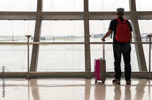 Backpacker man with backpack and carry luggage standing and looking through airport windows in airport, Tourist And Vacation Concept