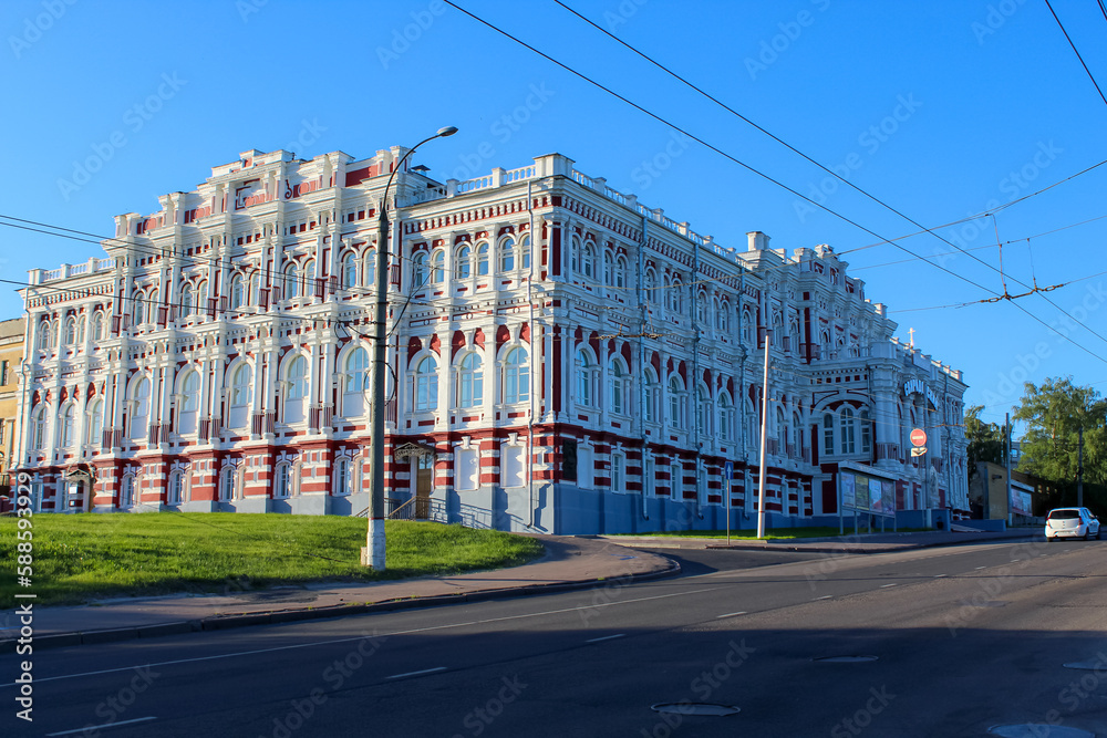Kursk city, Russia. The building of the Noble Assembly a three-story brick corner building in eclectic style, located in the historic centre of Kursk. The green lawn and blue sky create a frame.