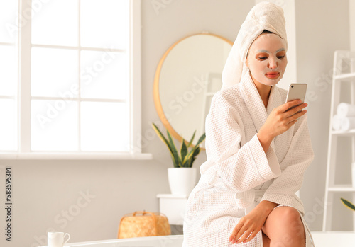 Young woman with sheet mask using mobile phone in bathroom