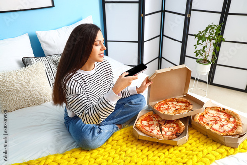Young woman with mobile phone taking picture of tasty pizza in bedroom