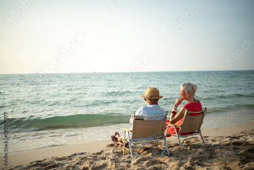 A couple of elderly sitting in chairs at the beach watching the sun and the sea on their summer vacation and they smile and enjoy their vacation.