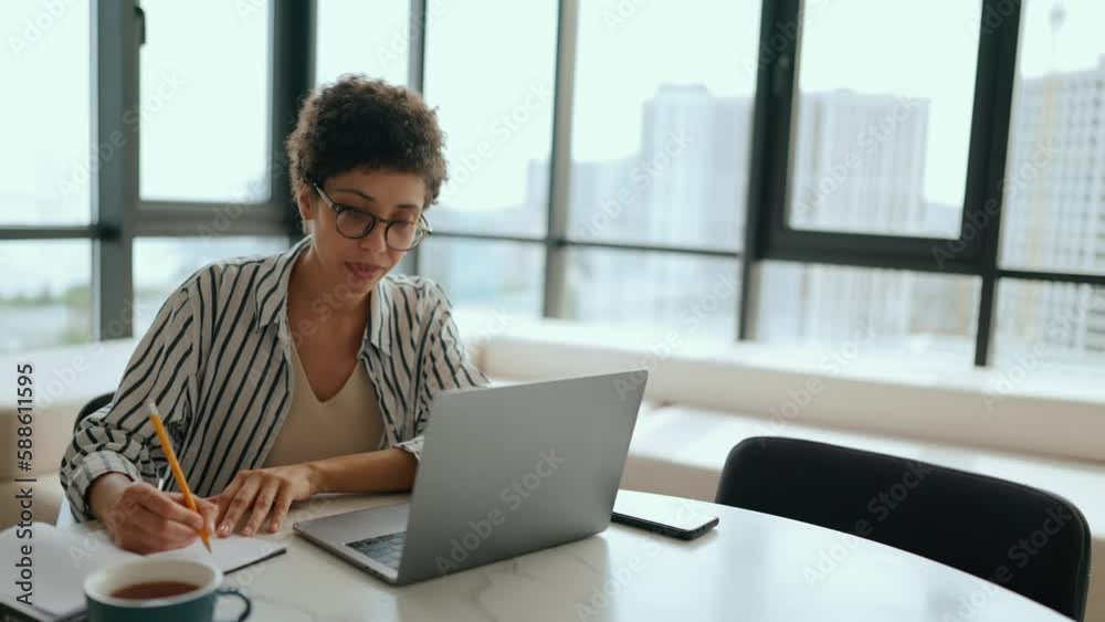 Concentrated Latin curly haired woman writing something in notepad from laptop at home Concentrated Latin curly haired woman writing something in notepad from laptop at home