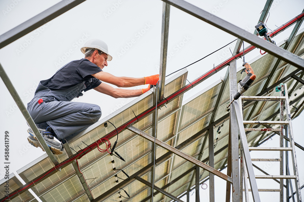 Man engineer installing photovoltaic solar panel system under white sky. Male workers in safety construction helmets mounting solar modules, panels and support structures of photovoltaic solar array.