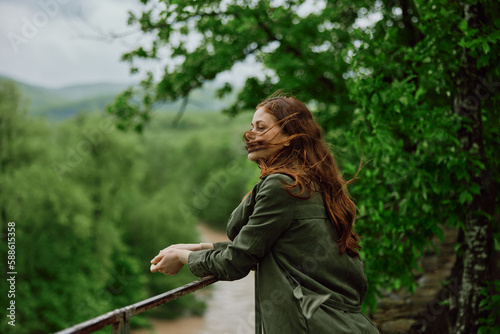 a beautiful woman with red hair blowing in the wind stands in a park overlooking a mountain river