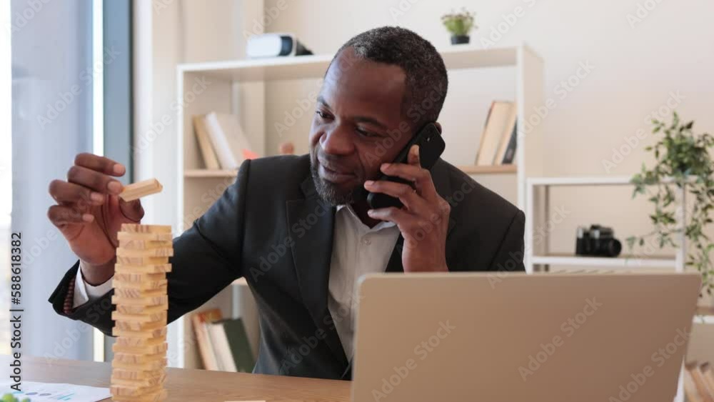 Happy multicultural man in business suit playing jenga while talking on smartphone at office desk with laptop. Cheerful entrepreneur reducing stress at work by enjoying board game during conversation.