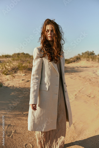 a red-haired woman stands in the desert in light clothes in windy weather looking at the camera