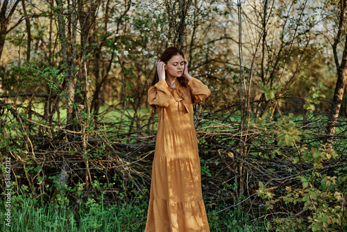 a beautiful woman walks in the shade near the trees, dressed in a long orange dress, enjoying the weather and the weekend, straightening her hair with her hands. The theme of privacy with nature