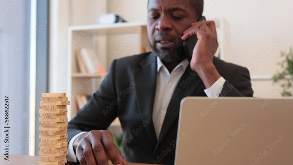 Happy multicultural man in business suit playing jenga while talking on smartphone at office desk with laptop. Cheerful entrepreneur reducing stress at work by enjoying board game during conversation.