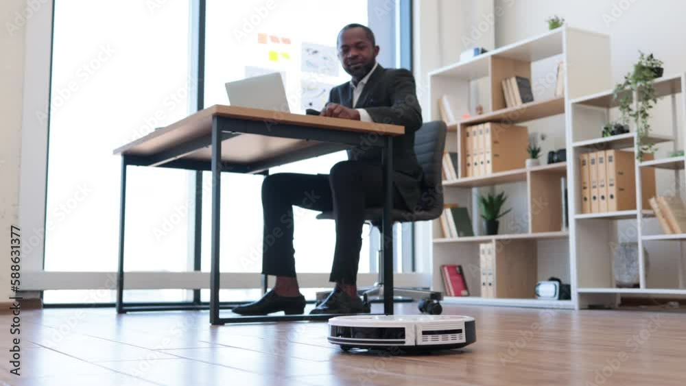 Remote-controlled robotic vacuum removing dust from laminate wooden floor while joyful african american businessman working on portable computer. Automatic cleaning improving workplace hygiene.
