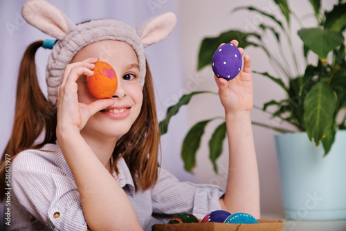 A positive playful girl in bunny ears holds colorful eggs in her hands in the home kitchen. Happy Easter. Easter background