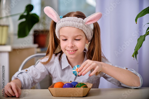 A positive playful girl in bunny ears holds colorful eggs in her hands in the home kitchen. Happy Easter. Easter background