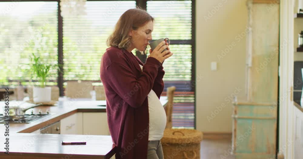 Caucasian pregnant woman standing in kitchen, touching belly and drinking tea Caucasian pregnant woman standing in kitchen, touching belly and drinking tea