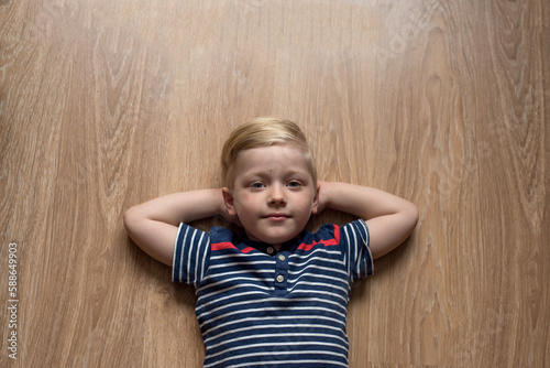 Young cute serious blond boy in stripped t-shirt is laying on the brown floor. Place for text