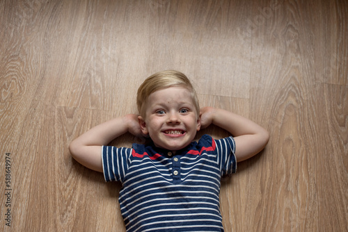 Young cute happy smiled blond boy in stripped t-shirt is laying on the brown floor. Place for text