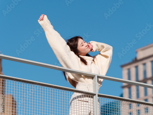 Beautiful young woman with black long hair in white skirt posing eyes closed with blue sky and city buildings background in sunny day. Emotions, people, beauty, travel and lifestyle concept.