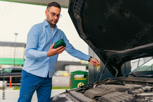 The man is standing in front of the raised hood, checking and changing the oil at the gas station