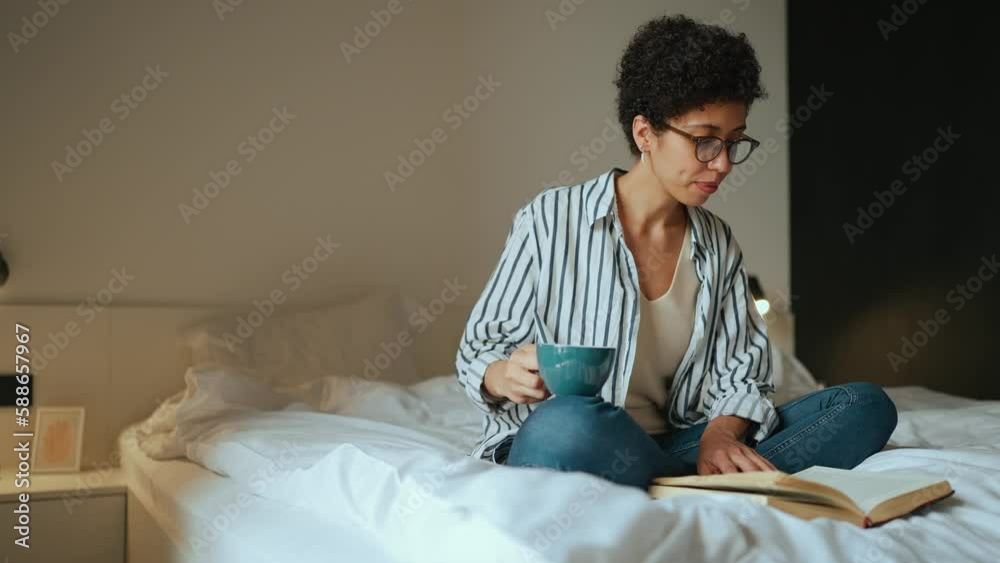 Smiling Hispanic curly haired woman reading a book with tea in hand and looking out the window on the bed Smiling Hispanic curly haired woman reading a book with tea in hand and looking out the window on the bed