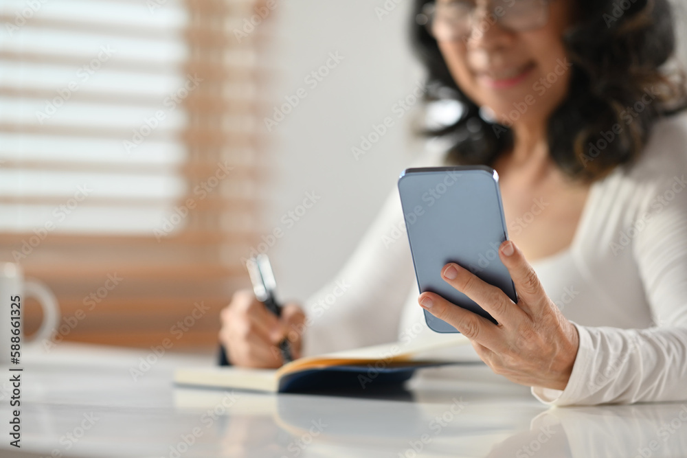 Selected focus on hand. Smiling senior woman checking working schedule plan on smart phone and making notes on notebook Selected focus on hand. Smiling senior woman checking working schedule plan on smart phone and making notes on notebook