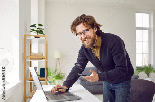 Successful satisfied Caucasian man entrepreneur uses laptop and mobile phone running own business fulfills customer orders via Internet dressed in casual clothes stands near with desk in home office