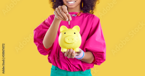 Happy kid saving up some money. Little child holding yellow piggy bank. Cheerful Afro American girl puts one coin inside her little piggybank. Cropped shot. Banner background. Finance concept