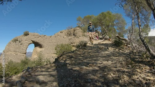 Man hike down on a mountain path  while a woman take photo of him. Stone arch and trees in background with a strong wind. Les pénitents in Alpes de Haute Provence, france.