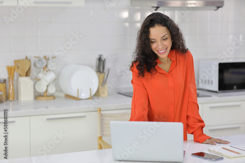 Smart and active latin woman standing and using internet on computer while using smart mobile phone with a cup of coffee in kitchen. Anywhere and work. Lifestyle with technology