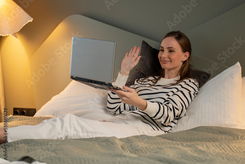 A cheerful young woman communicates with a friend via video call using a laptop while lying in bed in the bedroom. 