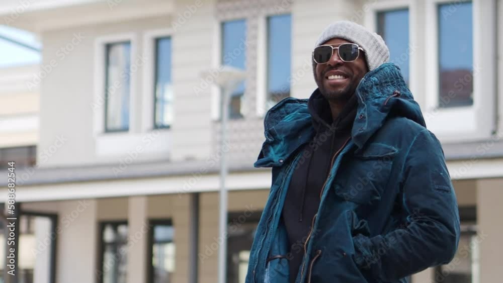 Portrait of handsome African American man standing and smiling in urban city, wearing stylish outfit parka coat, hoodie, knitted hat and sunglasses. street style fashion male model