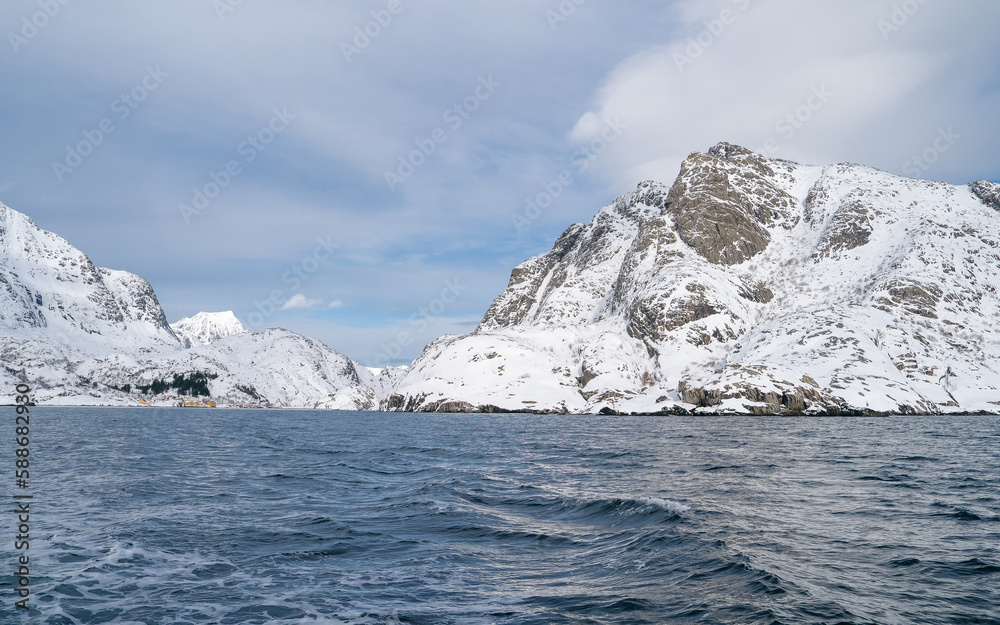 The calm waters reflect the rugged coastline and snow-capped peaks. In the foreground, the boat's ropes and rigging give a sense of movement and add a nautical element to the composition. Exploration