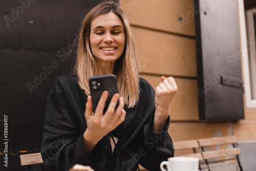 Joyful blonde woman playing video game on mobile phone, reading good news, celebrating winning lottery, feeling so happy and cheerful while making winner gesture, sitting at table with coffee cup.