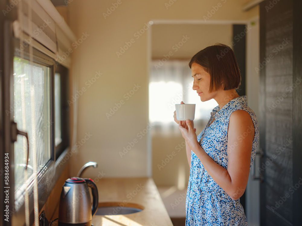 Young woman enjoying cup of coffee at morning, in her kitchen. Minimalism. Moving in. Living alone. Charming trailer house Young woman enjoying cup of coffee at morning, in her kitchen. Minimalism. Moving in. Living alone. Charming trailer house
