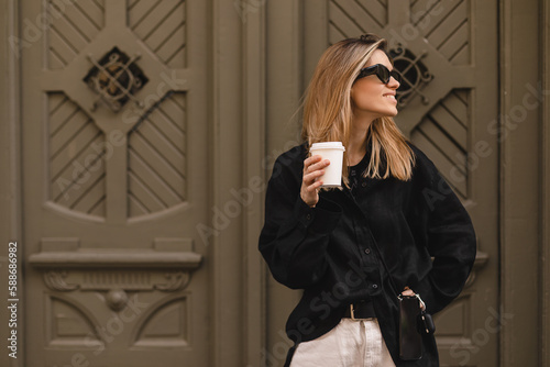 Happy beautiful stylish woman having good fashion clothes walking on street and holding coffee in cup takeaway with good mood. Girl in glasses look happy and look at side near green door.