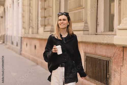 Smiling beautiful stylish woman having good fashion clothes walking on street and holding some coffee in cup takeaway with good summer mood. Girl look happy, turn around or spinning.