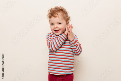 Portrait of funny little girl with blonde hair wearing casual striped turtleneck, having fun, playing, clapping hands, looking at camera, isolated on white background.