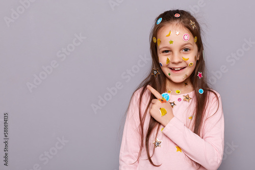 Image of happy joyful little brown haired little girl covered with stickers posing isolated over gray background, pointing finger aside at copy space for promotion.