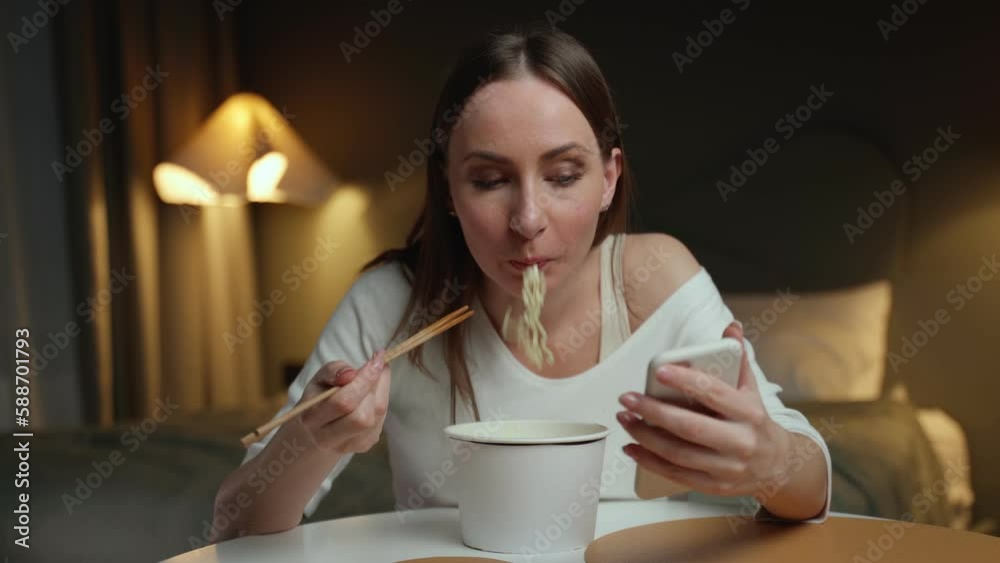 Busy woman uses a smartphone while having dinner in the bedroom. A student eats fast food while working on a new project in her home office Busy woman uses a smartphone while having dinner in the bedroom. A student eats fast food while working on a new project in her home office