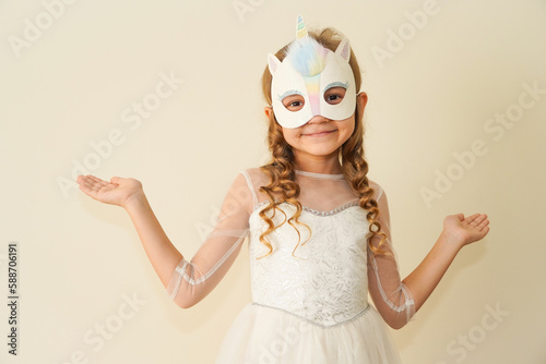  A child in a carnival mask of a unicorn, hand-made from glitter foamiran and faur fux. Little cute cheerful comical girl in the role of a unicorn, preparing for the carnival, masquerade, holidays.