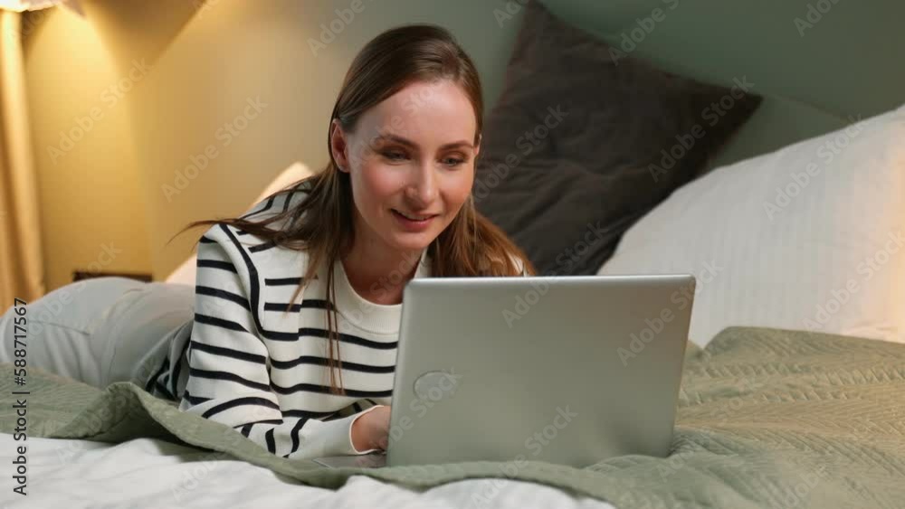 Beautiful young woman in a striped sweatshirt with a laptop is surfing the Internet, reading news on social networks while lying in bed. Beautiful young woman in a striped sweatshirt with a laptop is surfing the Internet, reading news on social networks while lying in bed.