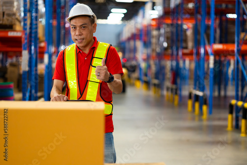 Warehouse staff worker wearing hardhat helmet standing by goods shelf working pulling a pallet truck in large warehouse factory. Asian engineer man