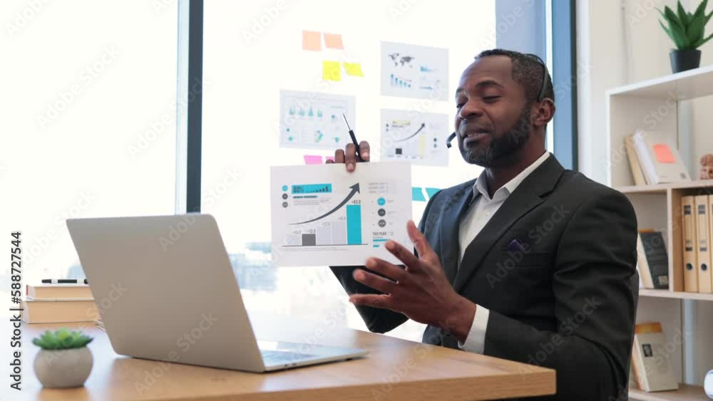 Formally dressed male entrepreneur sitting at workspace and having online meeting with colleagues on laptop. African american man in headset examining some documents holding in hands. Formally dressed male entrepreneur sitting at workspace and having online meeting with colleagues on laptop. African american man in headset examining some documents holding in hands.